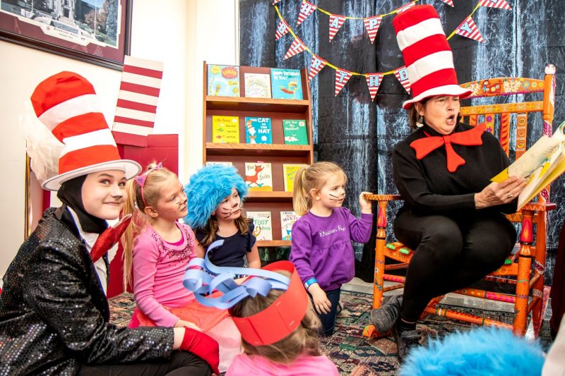A woman dressed all in black with a red-and-white striped top hat makes a face while she reads to a group of children from a brightly painted rocking chair.