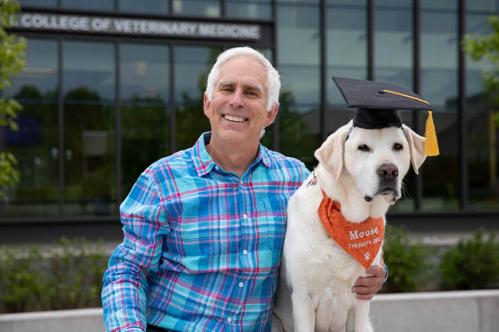 Moose, a yellow lab and therapy dog wearing a graduation cap, poses with owner/handler Trent Davis.