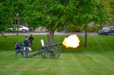 A cadet pulls the lanyard to fire the cannon on the Drillfield and smoke exits the barrel. Two other cadets stand watch nearby.