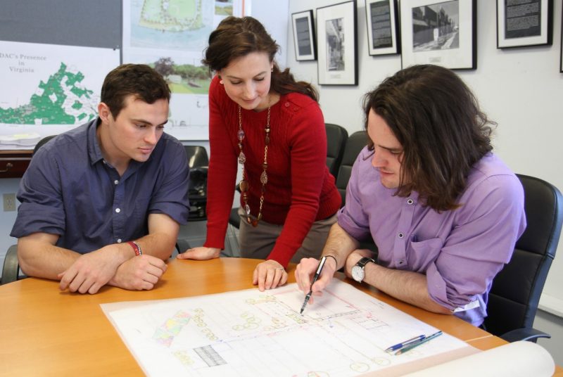 Two male college students and a woman gather around a landscape drawing in a conference room. 
