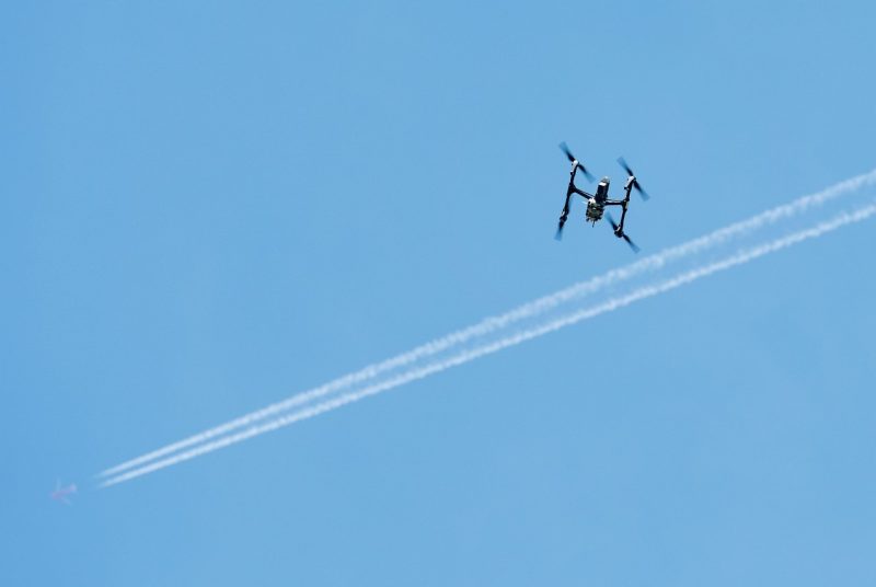 A drone is silhouetted against a blue sky with jet con trails behind it. 