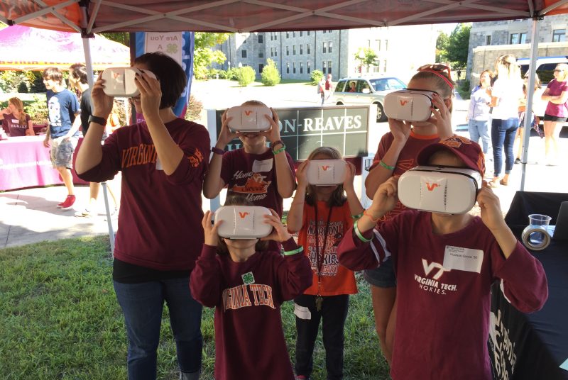 Several adults and children look through virtual reality goggles under a tent.