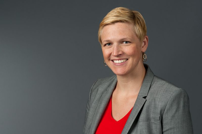 headshot of blond woman with short hair in gray jacket and red shirt