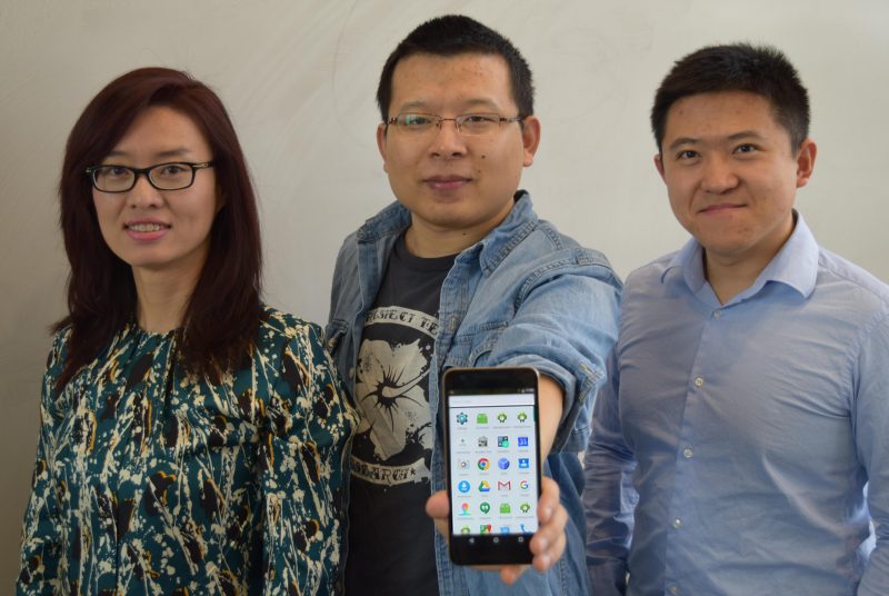 THree people stand in front of a white board and middle person holds Android phone out.