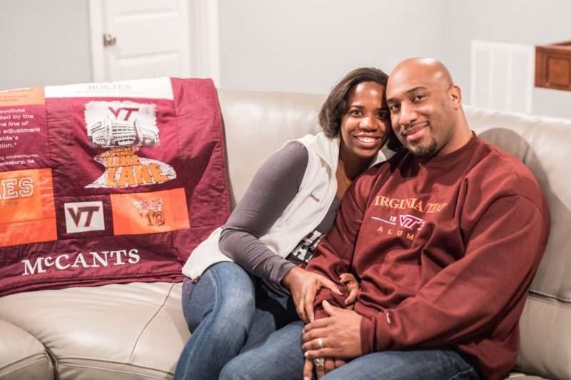 The young couple smiles and poses in their home, sitting on a couch with a blanket that is stiched with the last name "McCants" and made from orange and maroon shirts.