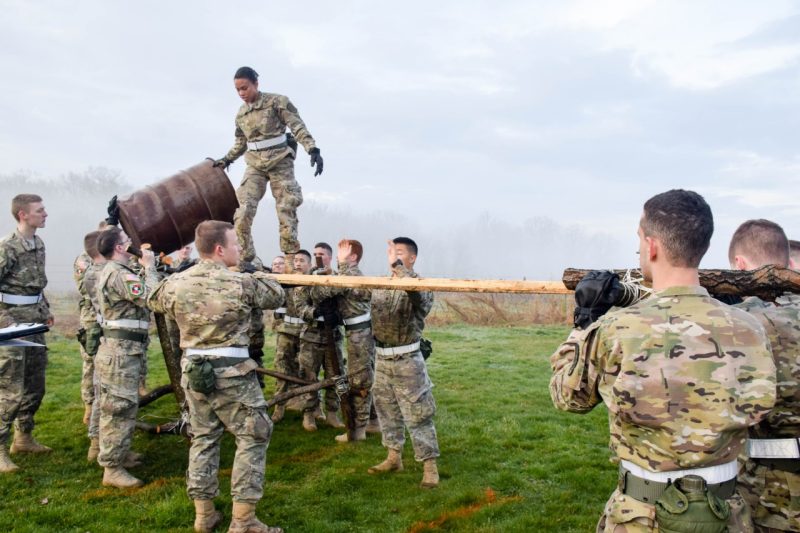 A cadet lifts a 50-gallon drum onto a makeshift bridge being supported in place by other cadets.