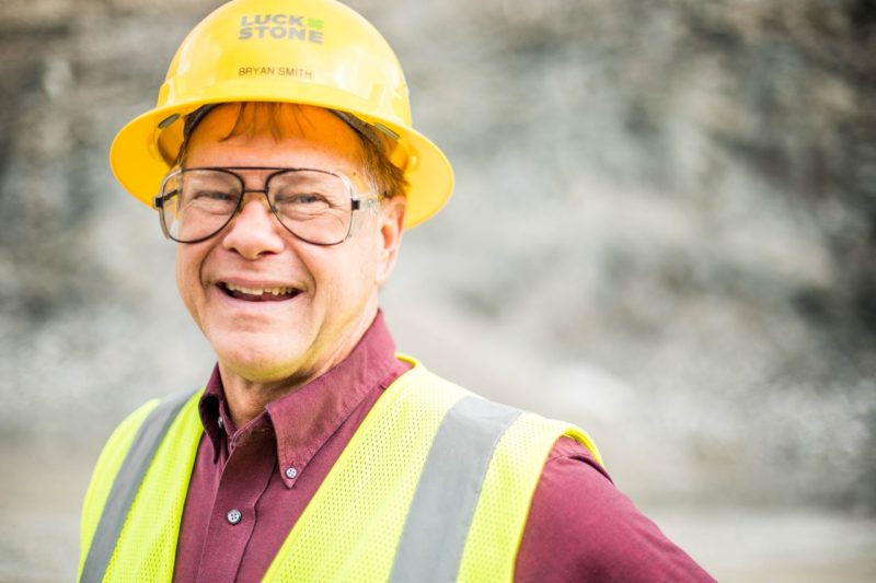 A portrait of a man standing inside a quarry wearing a yellow hard hat.
