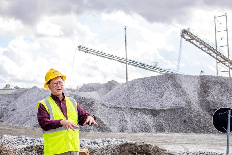 A man wearing a hard hat stands in front of large piles of aggregates, or small rocks, as more pour into the piles from overhead via large conveyor belts and machines.