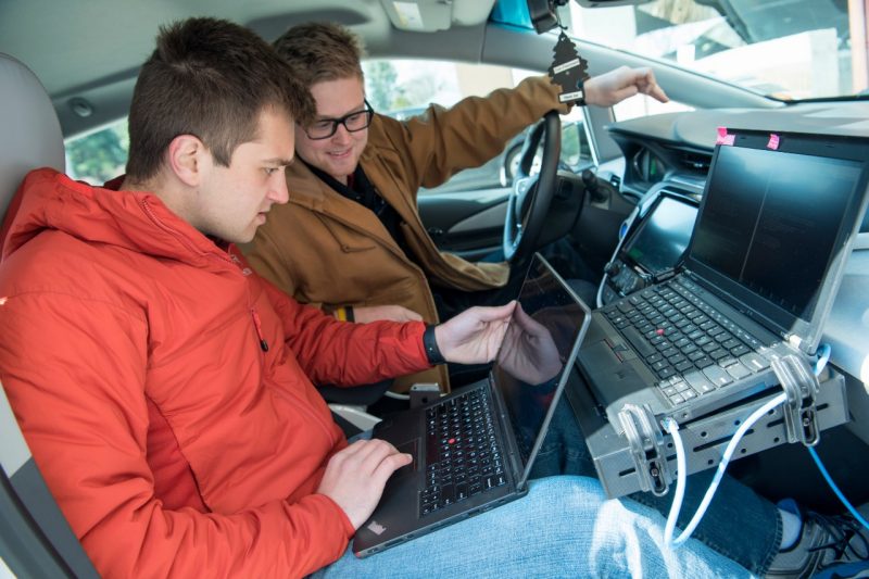 Two students sit inside of the vehicle and work from a laptop.