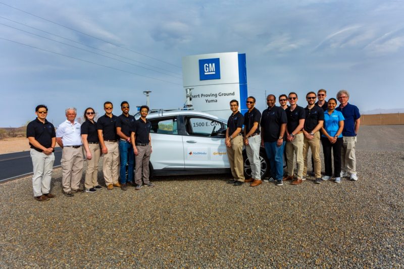 A team of people poses beside a white vehicle in the desert, in front of a sign that reads "GM Desert Proving Ground, Yuma."