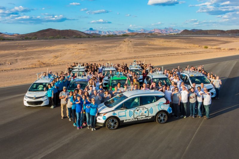 Dozens of people stand near eight small white vehicles and pose for a photo taken from above. In the background is desert landscape stretching on for miles.