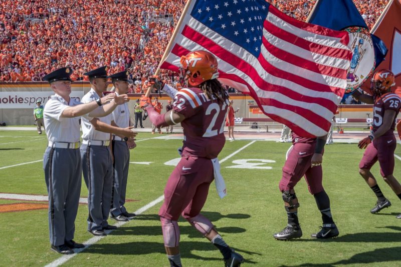 Cadet John Mitchell, at left, reaches for the American flag carried by a Hokie football player. To his right are Cadets Abdel Hammad and Paul Pechie. 