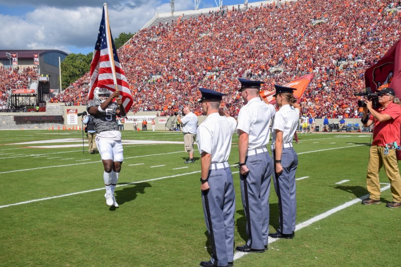 Three cadets stand at attention as a Hokie football player jogs toward them carrying the American flag.