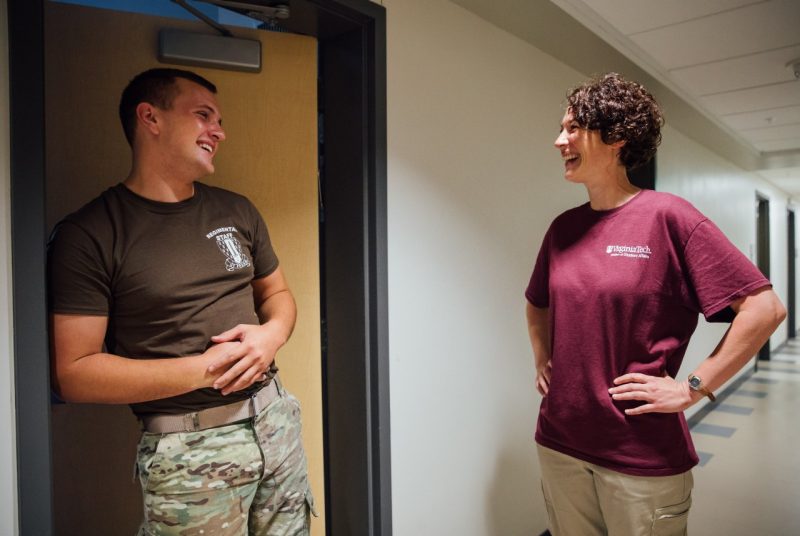 A cadet leans against the doorway of his residence hall room and smiles toward the woman he's talking to, who is standing in the hallway with her hands on her hips, also smiling