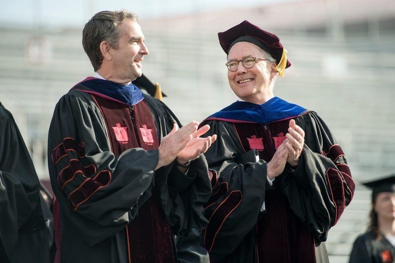 Two men wearing commencement regalia smile and clap.