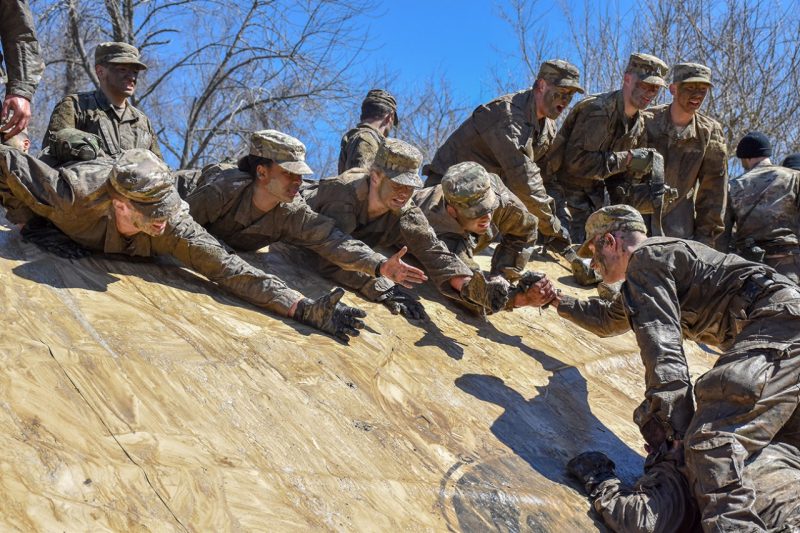 Cadets at the top of a wall reach out to pull a cadet over the top.