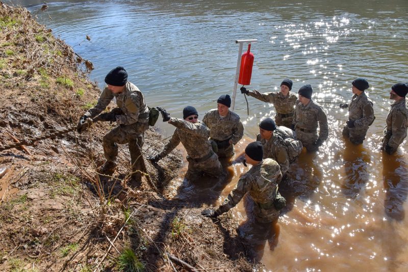 A cadet uses a rope to pull herself up a muddy river bank while other cadets wait in the water below.