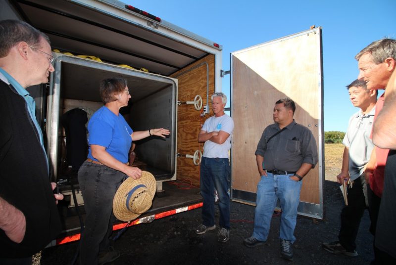 Six people — five men and one woman — stand in front of an open truck trailer containing a large metal box.