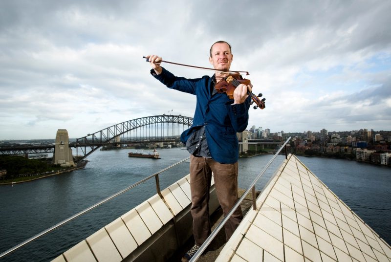 Violinist and Australian Chamber Orchestra artistic director Richard Tognetti plays his violin while standing on a bridge overlooking water.
