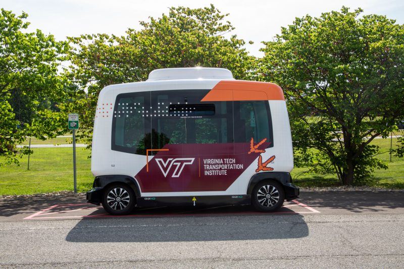 VTTI's automated shuttle is parked outside of the VTTI building. The vehicle is boxy and painted white, orange, and maroon.