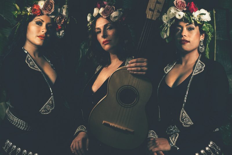 Three members of the band Flor de Toloache pose with instruments, wearing flower crowns.