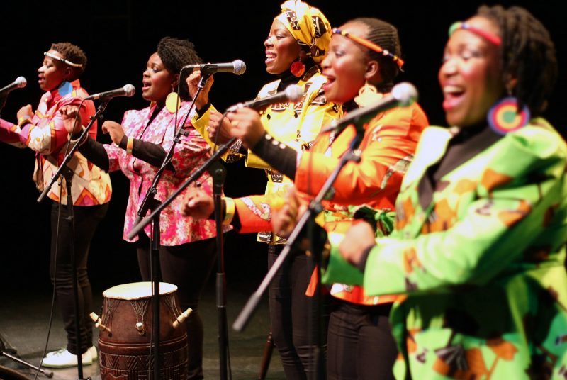 All female group Nobuntu are dressed in bright costumes and singing into microphones as they perform onstage.