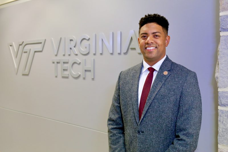 Photo of Kory Trott, standing in front of a silver "Virginia Tech" logo, which is mounted on the wall behind him. Photo credit: Alex Crookshanks for Virginia Tech.
