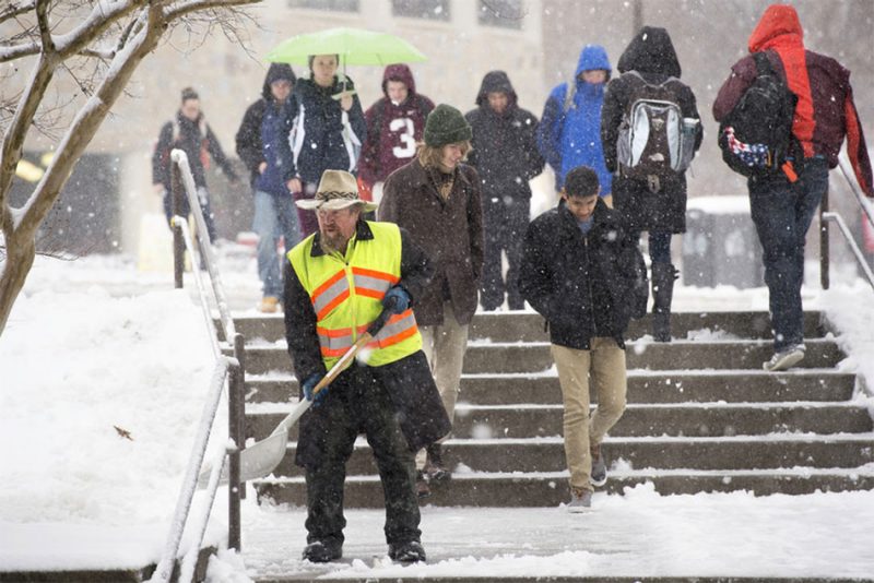 A facilities worker shovels a sidewalk as snow falls on an already blanketed ground. Students pass by.