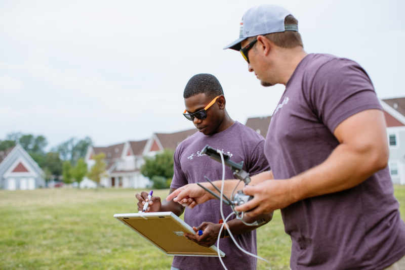 Morehouse undergraduate student Bryan Bloomfield and professor David Schmale record data during a field experiment at Grand Lake St Marys, Ohio. Bloomfield is holding a whiteboard with his left hand and hovering a marker using his right hand. Schmale is holding a drone remote controller in his left hand, while pointing at Bloomfield's whiteboard with his right hand. Photo courtesy Christina + David.  