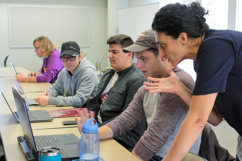 Professor talks closely with row of students working on laptop computers. 