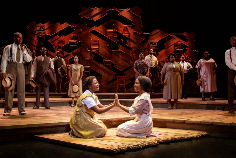 Two actresses kneel at the center of the stage, facing each other and holding up their hands, mirroring, while surrounded by other cast members in a stage scene from "The Color Purple."