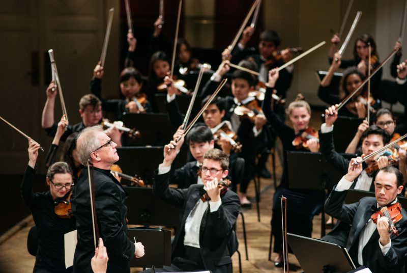 A conductor dramatically leads a group of string players, who all have their bows up in the air, onstage.