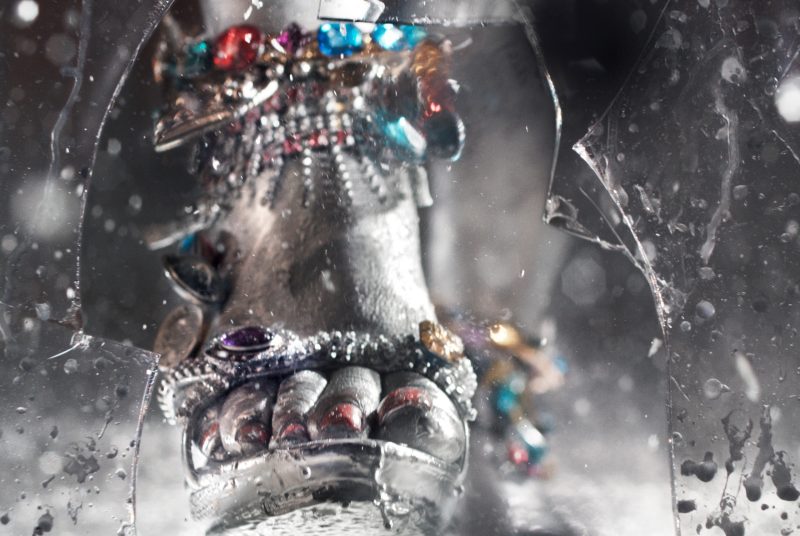A still image from a video created by artist Marilyn Minter of a woman's foot in a jeweled high heel sandal smashing through a piece of glass. 