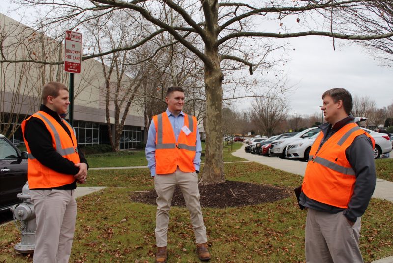 Three men stand outside an office building wearing orange reflective vests.