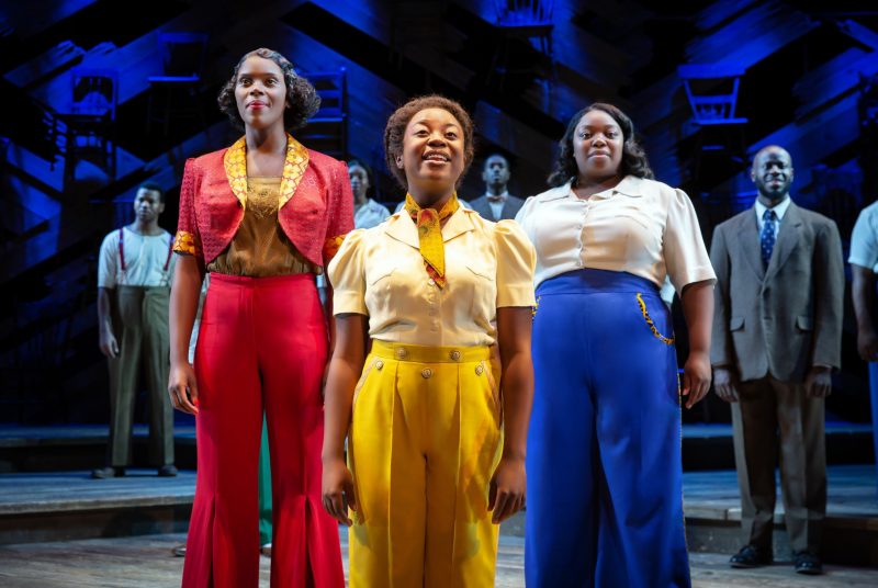 Three female performers in white shirts and brightly-colored pants take center stage during a performance of The Color Purple.