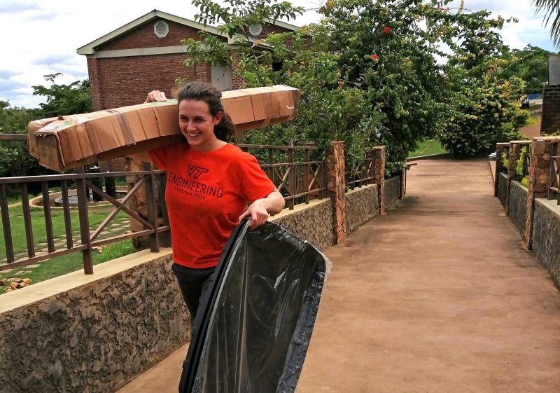 Brianna Friedman, a master’s student in mechanical engineering, is wearing an orange VT mechanical engineering shirt and walking across a small bridge while carrying a box on her shoulder and other equipment, aircraft, and supplies in her hand.