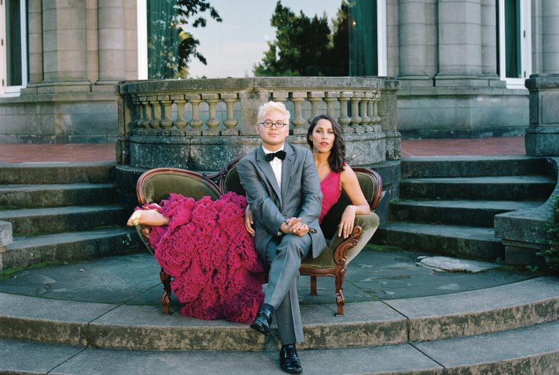 China Forbes and Thomas Lauderdale of Pink Martini pose on a lounge outside in front of pillars.
