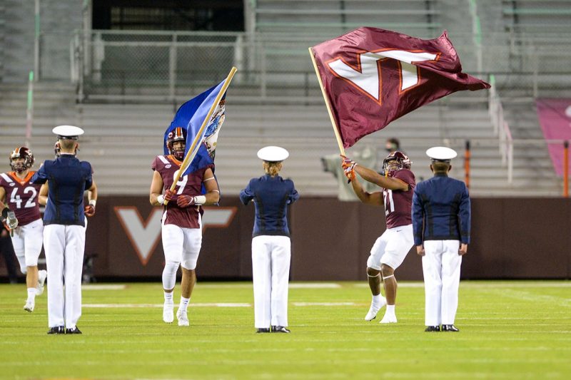 Three student cadets stand ready to receive flags carried by members of the Hokie football team. 