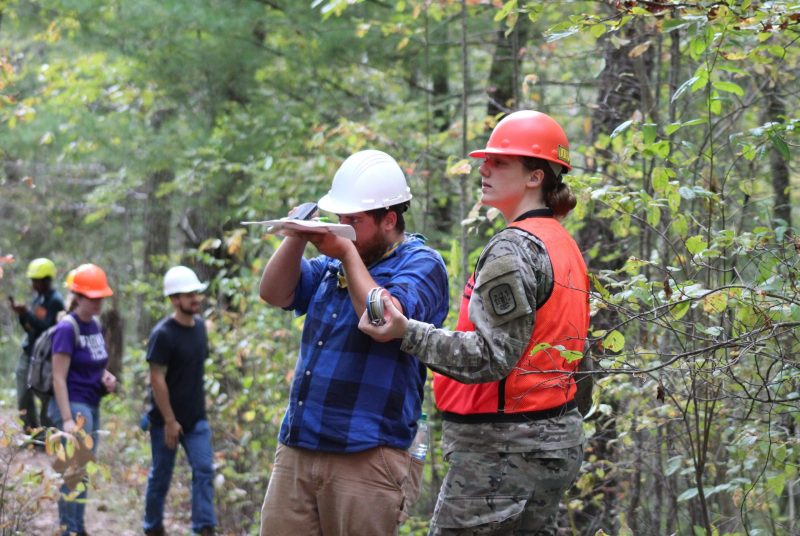 A young woman wearing fatigues, a reflective vest, and a hardhat stands in a forested area with several other people in the background.