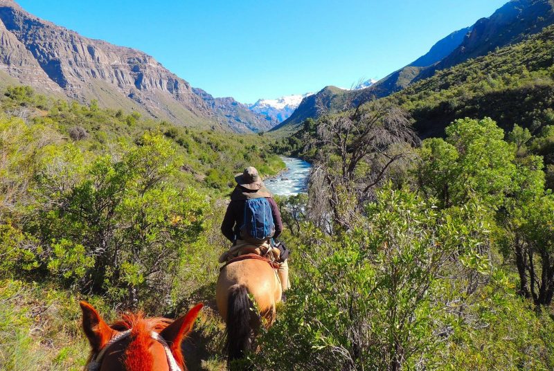 A person on horseback rides among shrubby vegetation into a wide mountain valley with a large creek running through it.