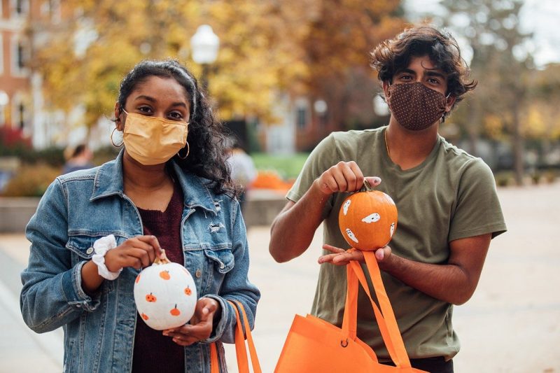 Two students, donning masks, pose with small painted pumpkins