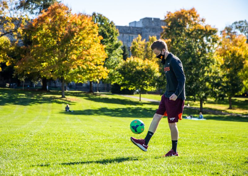 A student donning a mask kicks a soccer ball on the Drillfield