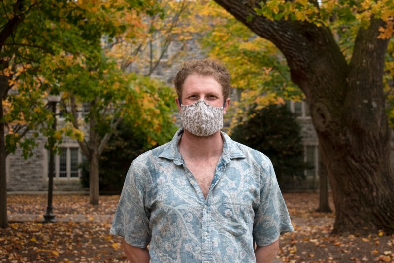 Virginia Tech College of Science Ph.D. student Ben Kligman  poses wearing a mask near Williams Hall on the Virginia Tech campus. Trees, with orange and yellow leaves, surround him. Photo by Steven Mackay.