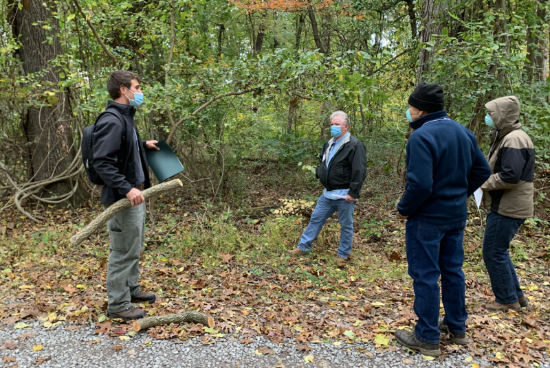 Four people standing on a gravel path in a wooded area. One faces the other three while holding a folder and a thick branch.