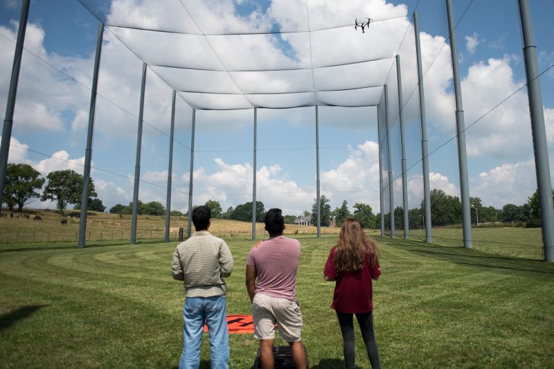 One female and two male students fly a drone in a netted enclosure. 