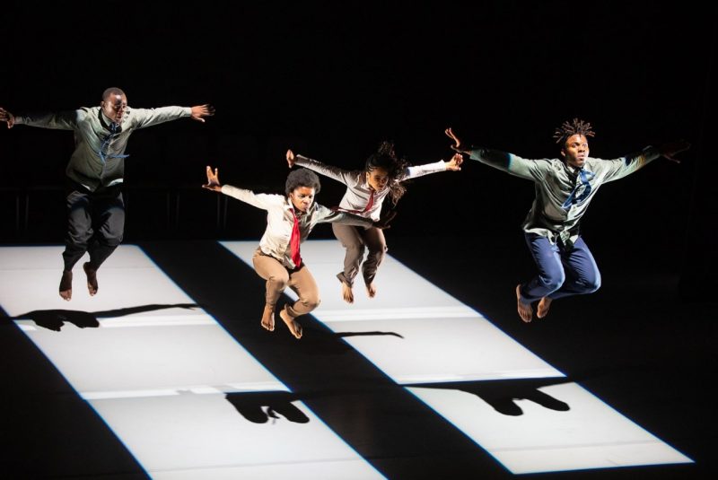 Four barefoot dancers dressed in pants, shirts, and ties are in the air-midjump, arms raised, with their shadows below.