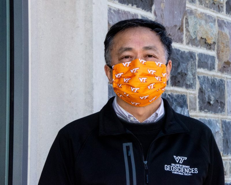 Xiao poses with orange Virginia Tech mask, and Geosciences jacket outside a Hokie Stone building.