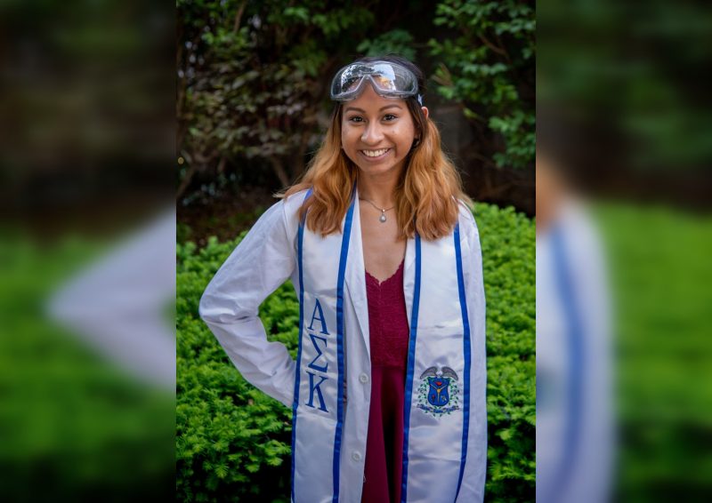 Amanda Ramirez stands with a hand on her hip wearing a white lab coat and lab goggles on the top of her head. She is also wearing a blue and white stole with the greek letters Alpha, Sigma, Kappa.