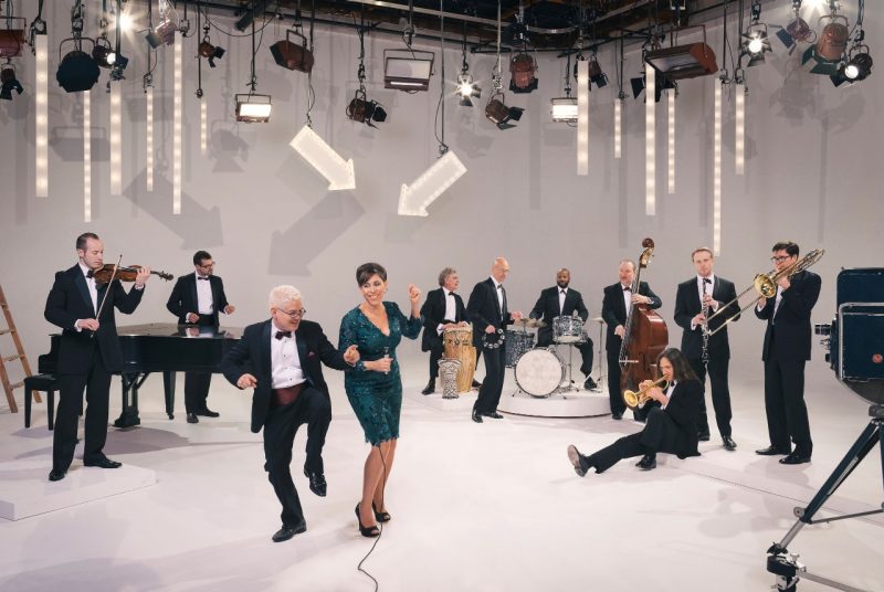 Eleven musicians with the band Pink Martini fill a clean, white room. A variety of stage lights hang overhead as they  hold their instruments - violins, drums, horns - while two members dance in the front.