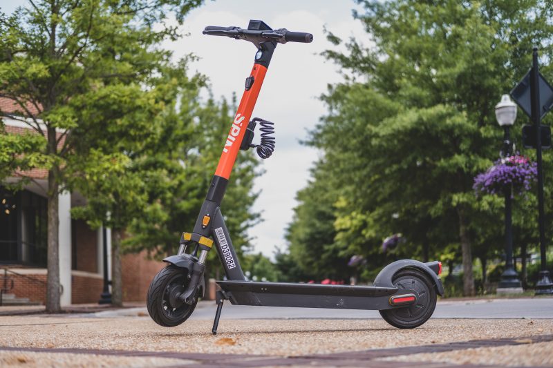 An orange and black electric scooter with the Spin logo is parked on a sidewalk surrounded by green trees